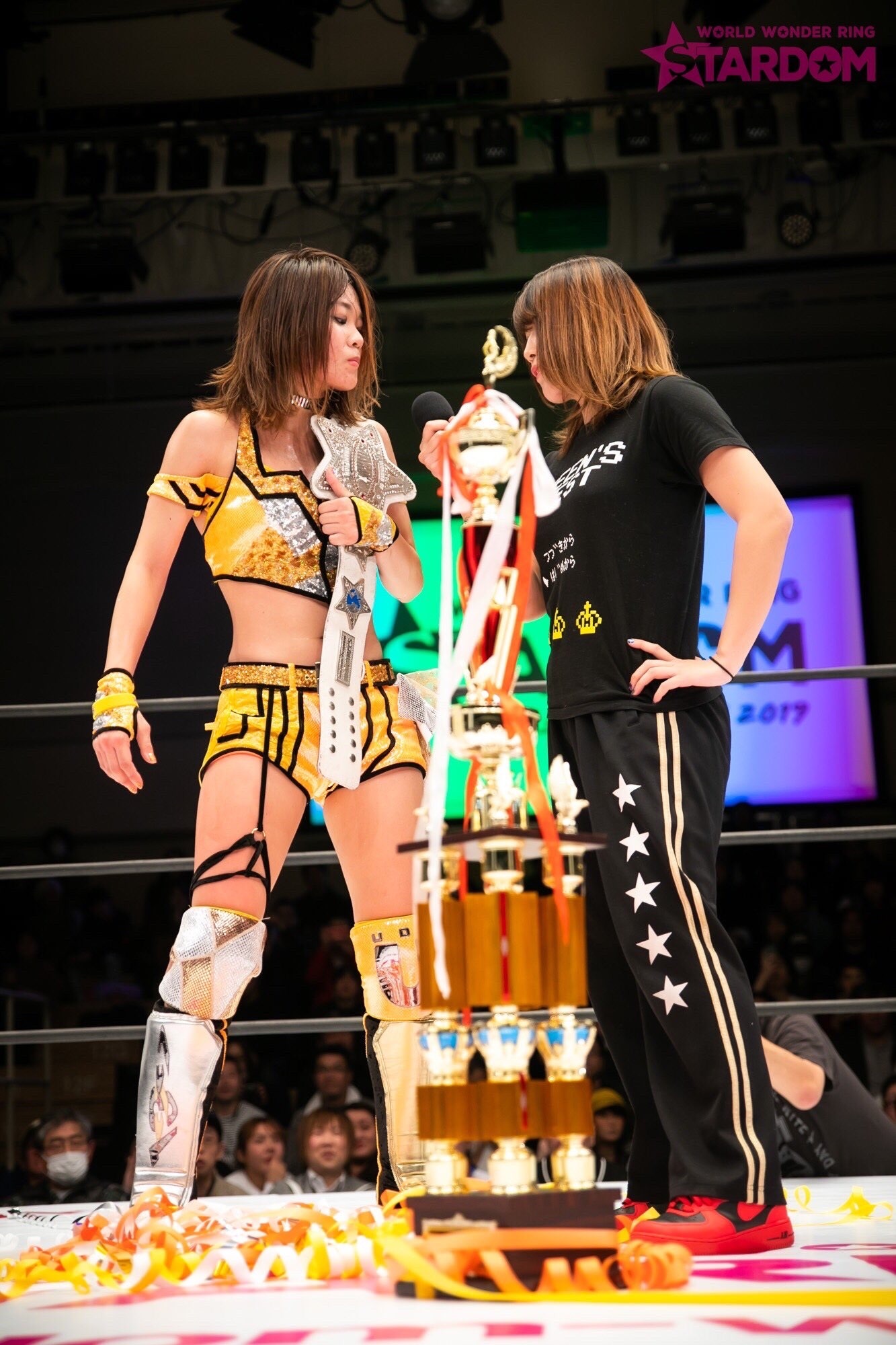 Arisa Hoshiki and Utami Hayashishita having a stare down behind a STARDOM trophy
